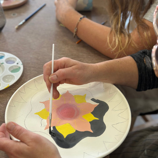 Person painting a ceramic plate with a floral design on a table.
